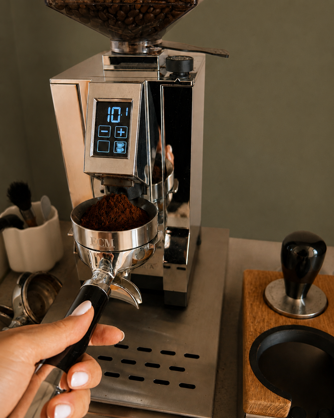 Hand hält den Siebträger mit frisch gemahlenem Kaffee unter der Mühle, dahinter die Espressomaschine im Morgenlicht