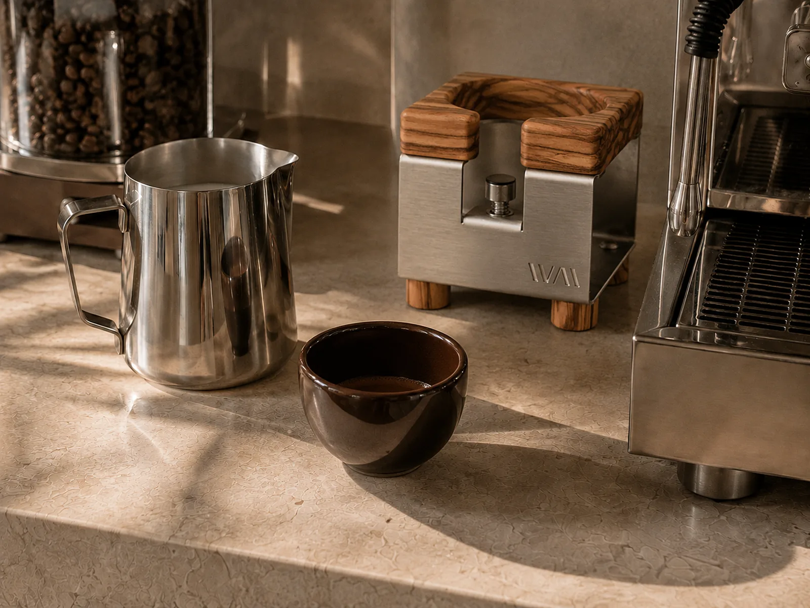 Milchkännchen, Tamperstation aus Edelstahl und Olivenholz und Cappuccinotasse raw cacao auf einer hellen Steinplatte.