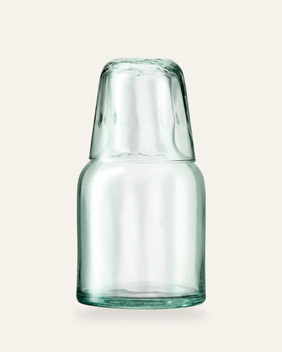 Wasserkaraffe aus recyceltem Glas mit aufgesetztem Trinkglas auf warmem Off-White.